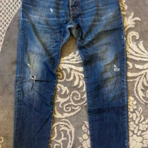 Dsquared2 jeans  - Snygga blå jeans från Dsquared2 med coola slitningar och tvättade detaljer. Klassisk femficksmodell med orange kontrastsömmar och Dsquared2-logga vid gylfen. Jeansen har en rak passform och är tillverkade i robust denim.