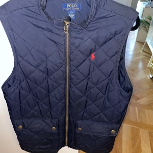 Quiltad blå väst från Polo Ralph Lauren - Mörkblå quiltad väst från Polo Ralph Lauren med dragkedja framtill, två fickor med tryckknappar och röd broderad logga på bröstet. Klassisk och stilren modell med hög krage, perfekt för lager-på-lager. Storlek L 14-16 men sitter mer som en S i herr. Jag är 175 och den sitter perfekt!