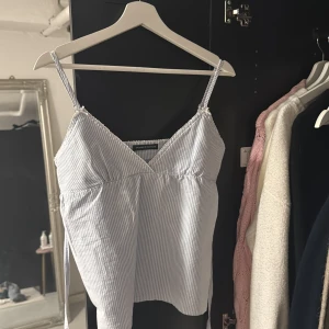 Randig blåvit linnetopp med knytband - Supersöt blåvit randig linnetopp från Brandy Melville med smala axelband och v-ringning. Toppen har knytband i midjan och är tillverkad i ett lätt bomullstyg, perfekt för varma dagar. Snygg och enkel att styla till jeans eller kjol. Finns ingen exakt storlek men är i xs/s