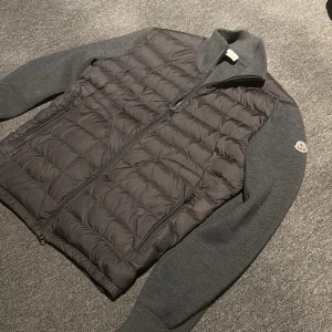 Moncler cardigan  - Snygg grå moncler cardigan från Moncler med quiltad framsida och stickade ärmar. Jackan har dragkedja framtill, två fickor med dragkedja och Moncler-logga på ärmen. Perfekt för kyliga dagar och ger en clean, modern look. Inget kvitto följer med men det är legit checkad så den är äkta, som ni även kan se på sista bilden på industri tagen 