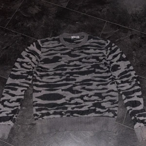 Grå/svart camo sweatshirt - Säljer en grå och svart stickad tröja med camouflagemönster. Tröjan har rund halsringning, ribbade muddar och är långärmad. Perfekt för dig som gillar streetstyle och vill sticka ut lite extra. Tröjan är köpt på zalando märket på tröjan är MO. Den är bara andvänd ett fåtal gånger. Skulle säga den är som en M-L. Priset är inte fast i sten🤙 