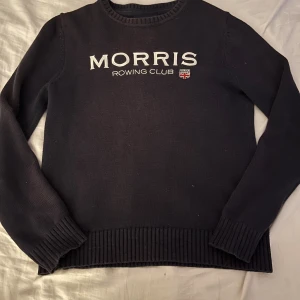 Mörkblå stickad tröja Morris Rowing Club - Snygg mörkblå stickad tröja från Morris Rowing Club med vit broderad logga och liten brittisk flagga på bröstet. Tröjan har rund hals, ribbade muddar och nederkant samt långärmad passform. Perfekt för dig som gillar klassisk stil med sportig touch.