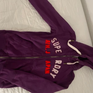 Lila hoodie från Superdry, Small - Lila hoodie från Superdry med bokstäver i vitt och rött framtill. Tröjan har huva med vita snören, dragkedja.  Snygg och bekväm passform( s i storlek men passar även xs). Jag köpte den från sellpy men den har fortfarande bra kvalitet och skulle inte säga att den har några större slitningar.🤍🤍