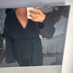 Svart stickad kofta med knytband - Mysig svart stickad kofta med breda ärmar och knytband i midjan. Koftan har en omlottmodell och är ribbstickad vilket ger en snygg struktur. Perfekt för lager-på-lager och chill dagar.