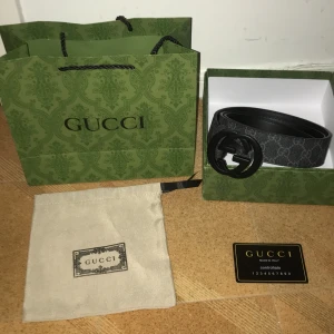 Svart och grått bälte med GG-spänne - Snyggt bälte i svart och grått med ett stort GG-spänne i matt svart. Bältet har ett diskret mönster och är tillverkat i skinn med textil på ovansidan. Perfekt för att lyfta din outfit med en lyxig touch.