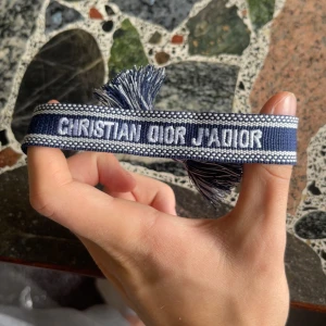 Blått Christian Dior J'Adior armband - Snyggt blått armband från Christian Dior med texten 'J'Adior' broderad i vitt. Armbandet har en bred, vävd design med fransar i ändarna och knytning. Perfekt accessoar för att lyfta din outfit med lite lyxig Dior-vibe.