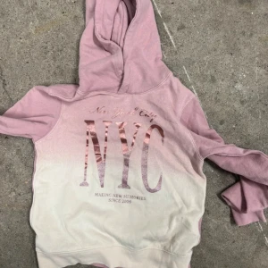 Rosa och vit hoodie med NYC-tryck från H&M - Säljer en hoodie från H&M i rosa och vitt med glittrigt NYC-tryck på bröstet. Tröjan har huva och mjukt material, perfekt för en avslappnad stil. Texten 'New York City' och 'Making new memories since 2006' står på framsidan. Ny och i storlek 146/152 ✨