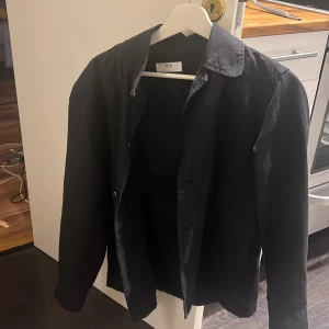 Svart overshirt från Uniqlo XS/S -  Tröjan är lite oversized vilket gör att den passar S Stilren svart overshirt från Uniqlo i storlek XS. Jackan har klassisk krage, knäppning framtill och raka ärmar. Perfekt för lager-på-lager och enkel att matcha med olika outfits. Materialet känns lätt och smidigt. 