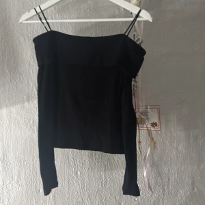 Svart långärmad topp från Stradivarius - Svart långärmad topp från Stradivarius med smala axelband och croppad passform. Toppen har en stilren, off shoulder, ribbat material som ger en snygg struktur. Perfekt för dig som gillar minimalistisk och trendig stil. Den kan vara lite kort i armarna 