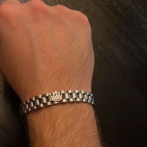 Silverfärgat armband med krona - Stilrent armband i silverfärgad metall med klassisk länkdesign och en snygg krona som detalj i mitten. Perfekt för dig som gillar minimalistiska accessoarer med en liten twist. Lätt att matcha med andra smycken och passar både killar och tjejer.