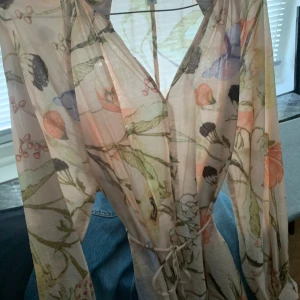 Blommig långärmad klänning med knyt - Superfin långärmad klänning i ljusa toner med blommigt mönster och v-ringning. Klänningen har ett smalt knytband i midjan och är tillverkad i ett lätt, luftigt material. Perfekt för dig som gillar romantiska och bohemiska vibes.