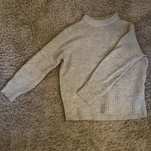 Grå stickad tröja från 157 - Mysig grå stickad tröja från 157 med högre ribbad krage och långa ärmar. Tröjan har en rak passform och breda ribbade muddar vid ärmslut och nederkant. Perfekt för lager-på-lager under kyliga dagar.