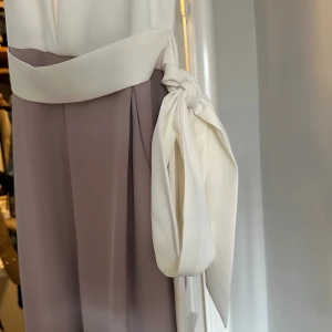 Jumpsuit från Phase Eight med volang - Supersnygg jumpsuit från Phase Eight med vit överdel, volangdetaljer vid axlarna och bred knytning i midjan. Kjoldelen är ljust lavendellila och har ett elegant fall. Perfekt för dig som vill ha en stilren och chic look.