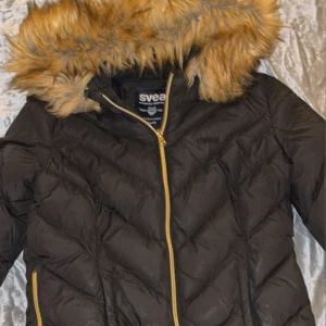 Svart pufferjacka från Svea med fuskpäls - Säljer en svart pufferjacka från Svea med snygg guldfärgad dragkedja och huva med fluffig fuskpäls. Jackan har quiltad design, två sidofickor med dragkedja och lång ärm. Perfekt för kalla dagar och har en cool streetstil.