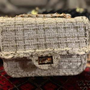Beige axelväska i tweed med kedja - Snygg beige axelväska i tweed med guldiga detaljer och rutigt mönster. Väskan har en kedja med inslag av vitt och guld, samt ett elegant lås framtill. Perfekt storlek för det viktigaste och ger en chic vibe till din outfit.
