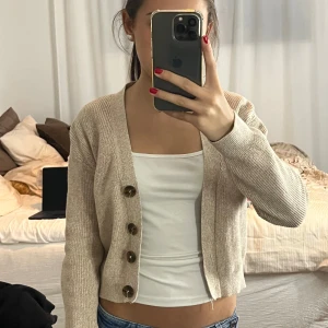 Beige stickad kofta med stora knappar - Superfin beige stickad kofta med stora bruna knappar framtill. Koftan är croppad och har lång ärm, perfekt att slänga över ett linne eller t-shirt. Enkel och stilren design som passar till jeans eller kjol.