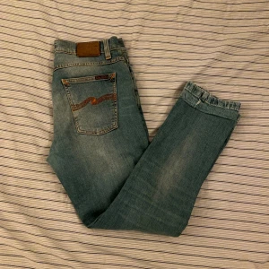 Nudie jeans - Säljer ett par klassiska blå nudie jeans med snyggt slitna detaljer och raka ben. Jeansen har fem fickor, bruna sömmar och logga på bakfickan. Perfekt passform och skön känsla i ett mjukt denimtyg. 30/30 - obs det är påsytt byxtygg längst ner (märks inte vid användning)