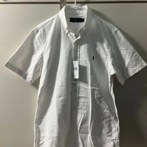 Vit kortärmad skjorta från Ralph Lauren - Vit skjorta från Ralph Lauren med korta ärmar och klassisk button-down krage. Skjortan har knappar framtill och den ikoniska blå Polo-loggan broderad på bröstet. Tillverkad i mjuk bomull, perfekt för en clean och stilren look.