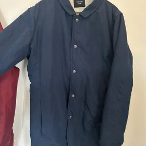 Quiltad blå jacka från Drop Dead - Mörkblå quiltad jacka från Drop Dead med stora vita broderade logotypen på ryggen. Jackan har tryckknappar framtill, raka ärmar och enkel krage. Perfekt för dig som vill sticka ut med en unik streetstyle-look.