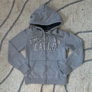 Grå hoodie från American Eagle - Grå hoodie från American Eagle med broderad logga framtill. Tröjan har dragkedja, huva med snörning och två fickor fram. Mjuk insida och ribbade muddar vid ärmslut och nederkant. Perfekt för chill dagar eller när du vill ha en avslappnad look.