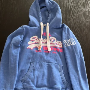 Blå Superdry hoodie med tryck - Blå hoodie från Superdry med vit och rosa logga framtill och siffran 28 på ärmen. Tröjan har huva med vita snören och en stor magficka. Perfekt för dig som gillar sportig och avslappnad stil. Kan lösas en snabb affär!