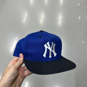 Blå New York Yankees keps - Snygg blå snapback-keps med klassisk NY-logga i vitt framtill. Skärmen är svart på ovansidan och grön undertill, och kepsen har justerbar plastrem bak. Perfekt för dig som vill ha sportig streetstyle och gillar baseball.