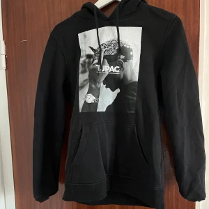 Svart Tupac hoodie från Mister Tee - Svart hoodie från Mister Tee med stor svartvit Tupac-print på bröstet. Klassisk känguruficka, snörning i huvan och mjukt material. Perfekt för dig som gillar streetstyle och vill ha en ikonisk look.