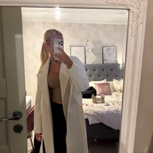 Vit lång kappa PULL& Bear - Säljer en stilren vit kappa med bred krage och stora fickor. Lite knopprig men går att få bort med en knoppringsmaskin. Perfekt för dig som vill ha en klassisk vibe i garderoben. Materialet känns mjukt och har en ullig känsla.