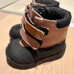 Kavat yxhult ash rose  vinterkängor st 20 - Säljer ett par Kavat yxhult storlek 20.  vinterkängor i rosa och svart med grov mönstrad sula för extra grepp. Skorna har två praktiska kardborreband och är fodrade med mjukt material för värme. Yttermaterialet är en kombination av skinn och syntet, perfekt för kalla dagar. Sparsamt använda som skor i den storleken ofta är. Skorna är nytvättade på bilderna så kommer i fint skick.