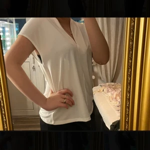 Vit v-ringad blus med kort ärm - Säljer en stilren vit blus med v-ringning och korta ärmar. Toppen har en lös passform och är tillverkad i ett mjukt, slätt material som faller snyggt. Perfekt för dig som gillar enkla och fräscha plagg med clean look. Aldrig använd!