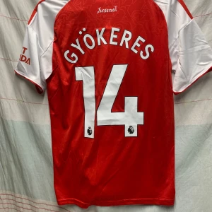 Arsenal Gyökeres #14 fotbollströja - Snygg Arsenal fotbollströja från Adidas med Gyökeres och nummer 14 på ryggen. Röd kropp med vita ärmar och detaljer, broderat Arsenal-emblem och Adidas-logga på bröstet. Materialet är lätt och andas, perfekt för match eller träning. Emirates Fly Better tryck på framsidan.