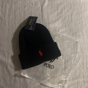 Svart ribbad mössa från Polo Ralph Lauren - Svart ribbstickad mössa från Polo Ralph Lauren med klassisk röd broderad logga framtill. Mössan har uppvikt kant och är tillverkad i mjukt material som håller dig varm. Perfekt accessoar för kyliga dagar och snygg till streetwear.