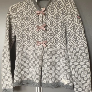 Gråvit kofta med rosetter Odd Molly - Superfin kofta från Odd Molly i grått och vitt med stickat mönster och tre ljusrosa rosetter framtill. Långärmad med broderad logga på ärmen och snygga detaljer vid kanterna. Perfekt för dig som gillar mysiga och unika plagg.