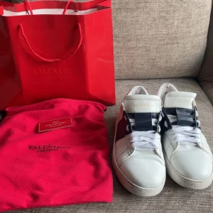 Valentino Garavani sneakers vit/blå - Snygga Valentino Garavani sneakers i vitt skinn med breda mörkblå detaljer över snörningen och coola nitar på hälen. Klassisk rund tå och platt sula, med vita skosnören och guldlogga på plösen. Perfekt för dig som gillar exklusiva streetwear-vibbar.