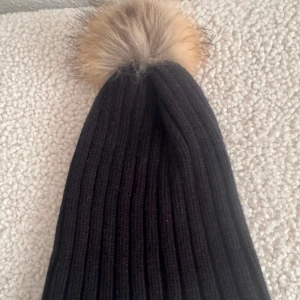 Svart ribbad mössa med pälsboll - Svart ribbstickad mössa med bred uppvikt kant och en fluffig pälsboll i beige och bruna toner på toppen. Perfekt för kalla dagar och ger en snygg touch till din vinterstil. Materialet är stickat och mössan har en klassisk form.