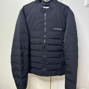 Svart tunn dunjacka från Acne Studios storlek 46 - Svart dunjacka från Acne Studios med minimalistisk design och diskret logga på bröstet. Jackan har quiltad struktur, rak passform och dragkedja framtill. Yttermaterialet är nylon och fodret består av 90% dun och 10% fjäder. Perfekt för kyliga dagar. Storlek 46