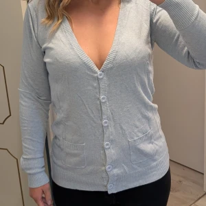 Ljusgrå kofta med knappar - Ljusgrå kofta med v-ringning och långa ärmar. Tröjan har ribbade muddar, två små fickor framtill och stängs med sex knappar. Materialet är mjukt. Enkel och clean stil som funkar till det mesta. Börjar få lite noppor se bild.