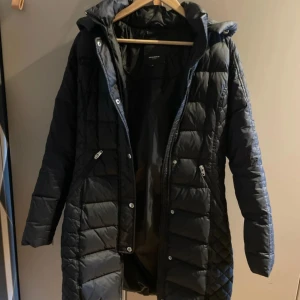 Svart lång dunjacka Rockandblue - Säljer en svart lång dunjacka från Rockandblue med huva, quiltade detaljer och dragkedjor. Jackan har snygga fickor med dragkedja och ett coolt märke på ärmen. Perfekt för kalla dagar och har en stilren look som funkar till allt.