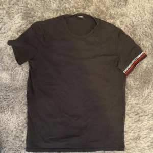 Svart t-shirt från Dsquared2 - Svart t-shirt från Dsquared2 med rund hals och korta ärmar. Ena ärmen har en snygg detalj med Dsquared2-logga och röd rand. T-shirten är i mjuk bomull och har en clean, stilren look som passar perfekt till jeans eller shorts.