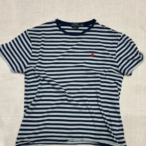 Randig t-shirt från Polo Ralph Lauren - Snygg randig t-shirt från Polo Ralph Lauren i marinblått och ljus grå med klassisk röd broderad logga på bröstet. T-shirten har rund halsringning och korta ärmar. Tillverkad i mjuk bomull för en skön känsla.