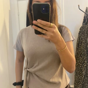 Beige kortärmad topp med knyt - Säljer en beige kortärmad topp med snygg knytning i sidan. Toppen har rund hals och är i ett mjukt, stickat material som känns skönt mot huden. Perfekt att styla med jeans eller kjol för en chill och trendig look.