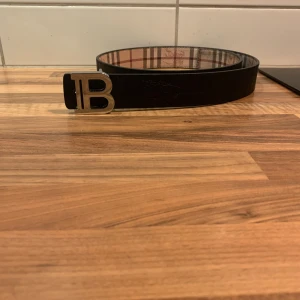 Burberry bälte med rutigt mönster - Snyggt Burberry bälte med klassiskt rutigt mönster i beige, brun och röd. Bältet har en stor metallspänne med B-logga och är tillverkat i skinn. Perfekt för dig som vill ha en stilren accessoar med lyxig känsla.