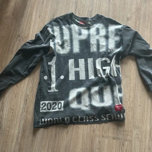 Supreme långärmad tröja med tryck - Supreme långärmad tröja i mörkgrått med stora vita tryck och textdetaljer över hela framsidan. Crewneck-modell i 100% bomull, perfekt för dig som gillar streetwear och statement-plagg. Röd Supreme-label nertill.