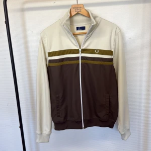 Randig tracktop från Fred Perry - Snygg tracktop från Fred Perry i storlek L. Jackan har en cool färgblockad design med beige, bruna och olivgröna ränder, samt vit dragkedja och ribbad krage. Ikonisk Fred Perry-logga broderad på bröstet. Perfekt för dig som gillar sportig och retro stil.