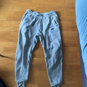 Grå mjukisbyxor från Nike med dragkedja - Grå mjukisbyxor från Nike med svart logga på låret och svarta snören i midjan. Byxorna har elastisk midja, ribbade muddar vid bensluten och praktiska fickor med dragkedja. Perfekta för chill eller träning.