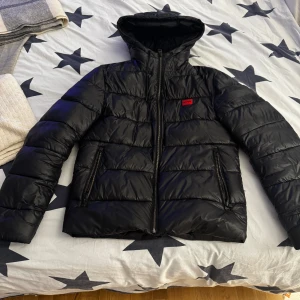 Svart pufferjacka från Hugo - Svart pufferjacka från Hugo med huva och rödmärkt logga på bröstet. Jackan har två fickor med dragkedja, quiltad design och hel dragkedja framtill. Perfekt för kalla dagar och riktigt snygg till jeans.