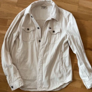 Overshirt från tiger of sweden - Snygg beige overshirt från Tiger of Sweden Jeans i storlek L. Skjortan har två bröstfickor med knappar, klassisk krage och långa ärmar med knappslå. Tillverkad i mjuk bomull som ger en chill och stilren vibe. Perfekt att slänga över en t-shirt för en clean look.