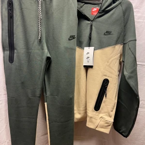 Nike hoodie i grönt och beige - Nike hoodie i tvåfärgad design med grön ovandel och beige nedre del. Tröjan har huva, dragkedja framtill och svarta detaljer på fickorna. Tillverkad i mjuk bomullsblandning, perfekt för chill eller träning. Klassisk Nike-logga på bröstet och sportig look.