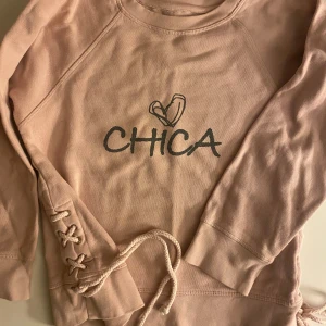 Rosa sweatshirt CHICA med snörning - Säljer en ljusrosa sweatshirt från CHICA med tryck framtill och cool snörningsdetalj på sidorna. Tröjan har rund halsringning och långa ärmar, perfekt. STORLEK 10År barn