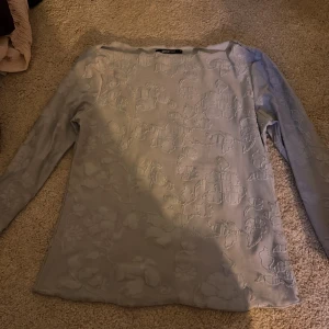 Ljusgrå blommig topp från Gina Tricot - Supersnygg ljusgrå topp från Gina Tricot med diskret blommigt mönster i tyget. Långärmad och i mjukt material som känns skönt mot huden. Perfekt att styla med jeans eller kjol för en trendig look.